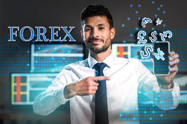 Gülümseyen iki ırklı tüccar akıllı telefonu işaret ederek Forex mektuplarının ve para işaretlerinin yakınındaki akıllı telefonu işaret ediyor. 