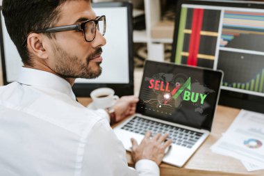 selective focus of bi-racial trader sitting near laptop with sell and buy letters clipart