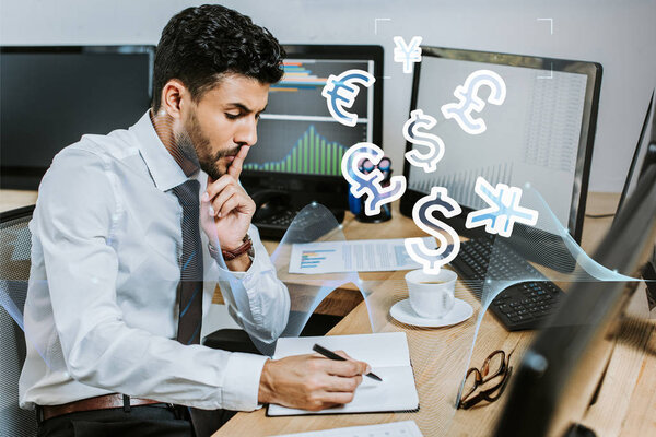 pensive bi-racial trader writing in notebook and sitting at table near money signs