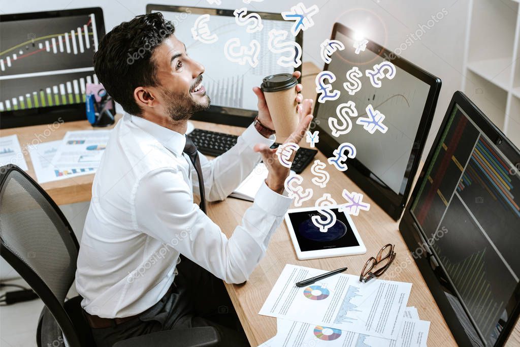 happy bi-racial trader holding paper cup and sitting near computers and money signs