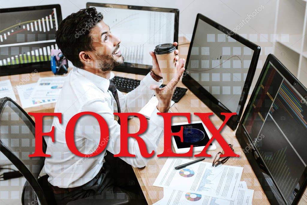 happy bi-racial trader holding paper cup and sitting near computers and forex letters