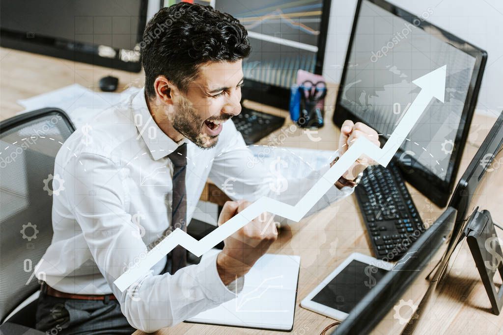 happy bi-racial trader showing yes gesture near computers and raising graphs 