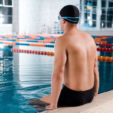 shirtless sportsman in swimming cap sitting near swimming pool  clipart