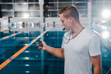 trainer in white t-shirt looking at timer while holding whistle in mouth  clipart