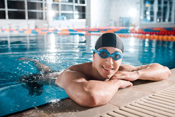happy swimmer in goggles and swimming cap looking at camera in swimming pool