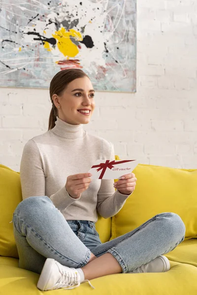 Beautiful Smiling Woman Holding Gift Voucher Looking Away Sofa Home — ストック写真