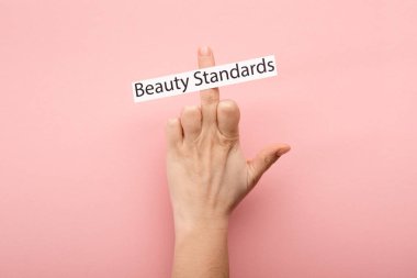 cropped view of woman showing middle finger and card with beauty standards lettering on pink background  clipart