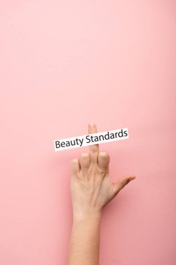 cropped view of woman showing middle finger and card with beauty standards lettering on pink background  clipart