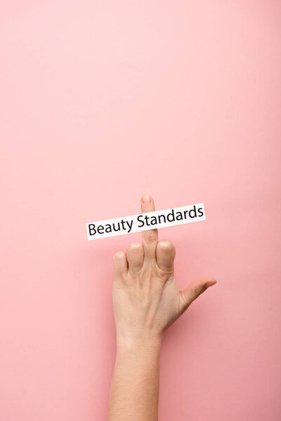 cropped view of woman showing middle finger and card with beauty standards lettering on pink background 