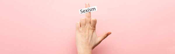 panoramic shot of woman showing middle finger and card with sexism lettering on pink background 
