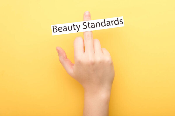 cropped view of woman showing middle finger and card with beauty standards lettering on yellow background 
