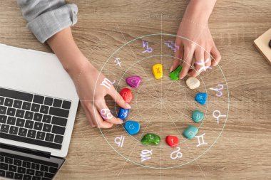 Cropped view of woman holding stones with zodiac signs beside laptop and illustration clipart