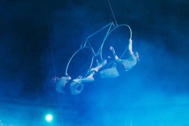 KYIV, UKRAINE - NOVEMBER 1, 2019: Low angle view of air gymnasts in smoke performing with rings in circus  clipart