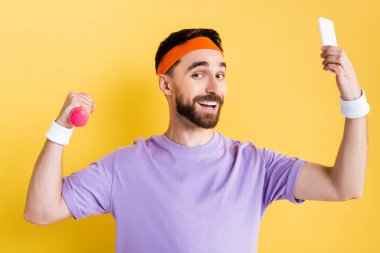 Neşeli sakallı sporcu selfie çekiyor ve sarı bir dambıl tutuyor.