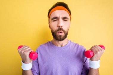 Sakallı sporcu, sarı renkli küçük dambıllarla egzersiz yaparken nefes alıyor.