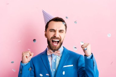 excited businessman in party cap celebrating triumph near falling confetti on pink  clipart