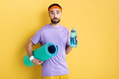  Sakallı adam elinde spor şişesi ve jimnastik paspası tutuyor.