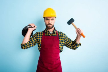 angry bearded workman holding hammer and dumbbell on blue  clipart