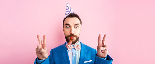 panoramic shot of businessman in party cap holding party blower in mouth and showing peace sign isolated on pink 