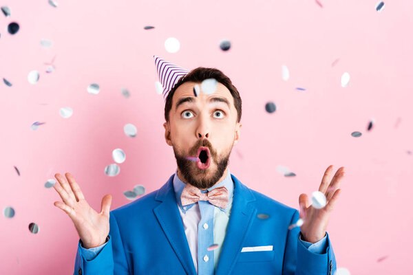 selective focus of surprised businessman looking at falling confetti on pink 