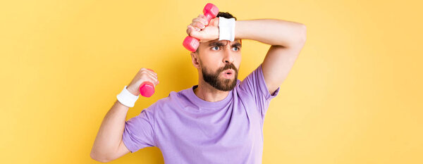 panoramic shot of tired bearded sportsman holding small dumbbells isolated on yellow