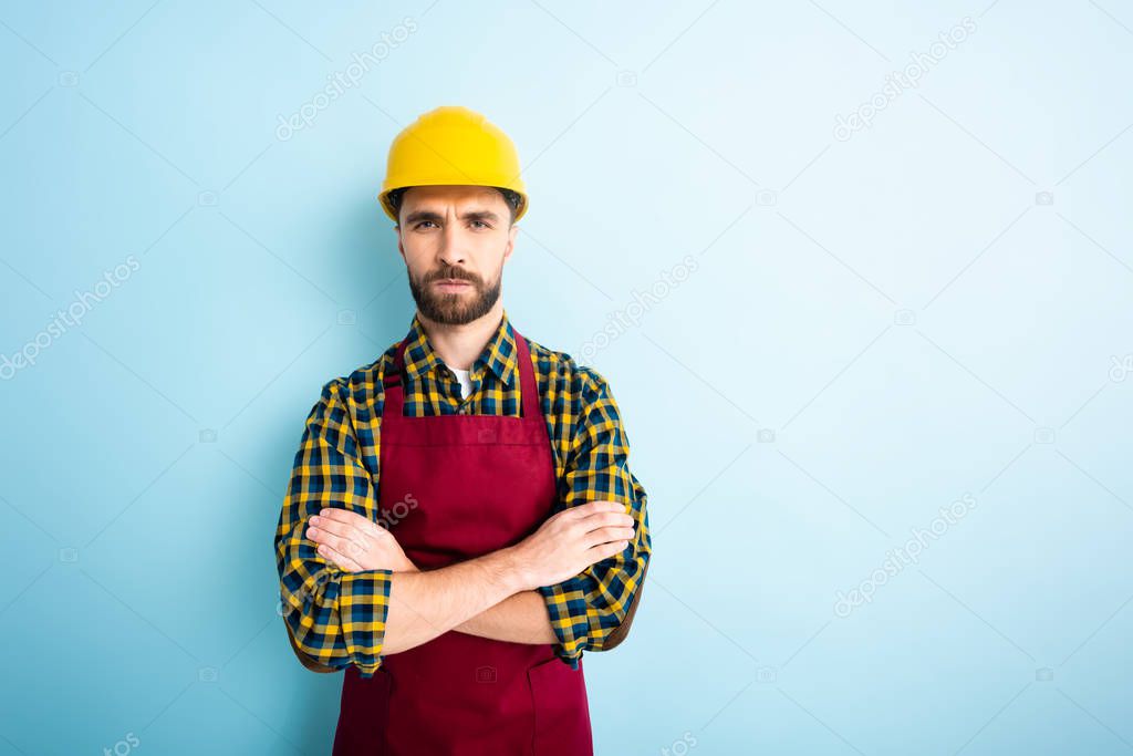 offended workman in safety helmet standing with crossed arms on blue 