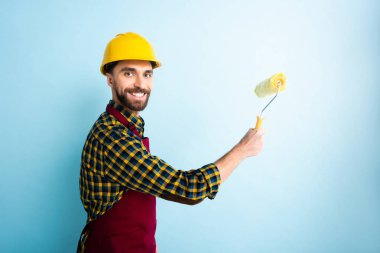 Mavi kasklı neşeli işçi elinde boya silindiri tutuyor. 