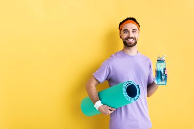 Pozitif sakallı adam elinde spor şişesi ve spor paspası tutuyor.