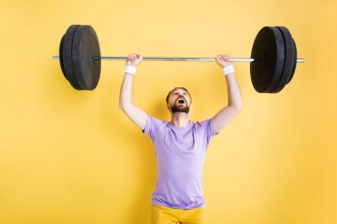 surprised bearded sportsman looking at barbell while working out on yellow clipart