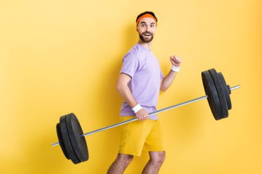 happy sportsman holding heavy barbell while walking on yellow clipart