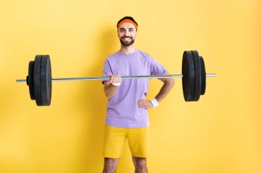 cheerful sportsman standing with hand on hip and holding barbell on yellow clipart
