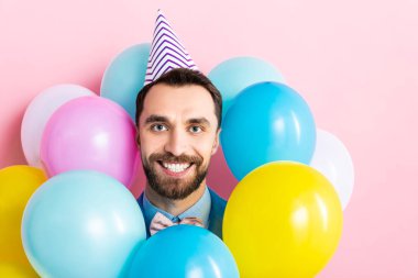 Parti şapkalı neşeli sakallı adam pembe üzerinde izole edilmiş renkli balonların yanında 