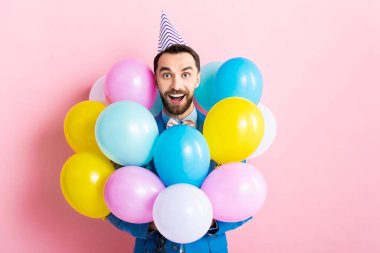 Parti şapkalı sakallı adam pembe renkli balonların yanında gülümsüyor. 