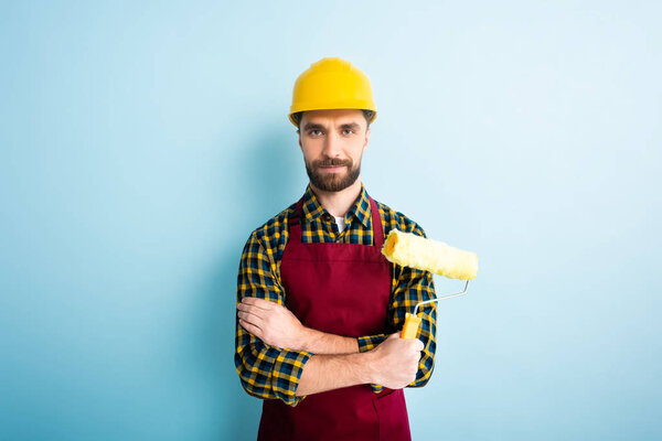 positive worker in safety helmet holding paint roller on blue 