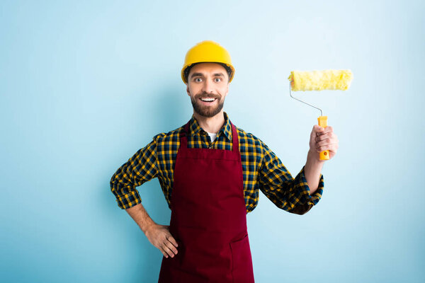 positive worker in safety helmet holding paint roller and standing with hand on hip on blue 