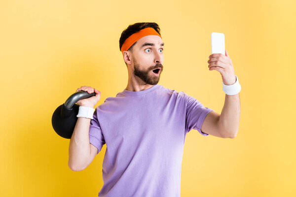 shocked sportsman working out with heavy dumbbell and taking selfie isolated on yellow