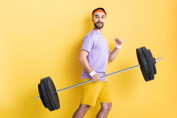happy sportsman holding heavy barbell while walking on yellow