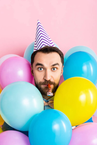 bearded man in party cap blowing party blower near colorful balloons isolated on pink 