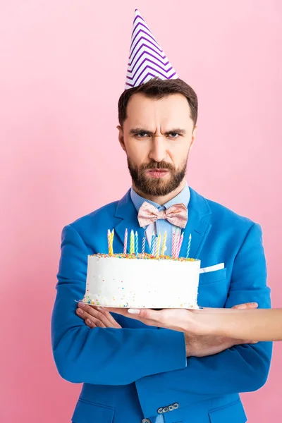 ピンクで隔絶された腕を持つ怒り狂った男の近くに誕生日ケーキを保持する女性のクロップドビュー — ストック写真