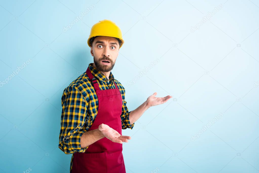 confused workman in safety helmet showing shrug gesture on blue 