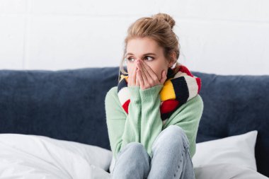 young ill woman in sweater and scarf sitting on bed  clipart