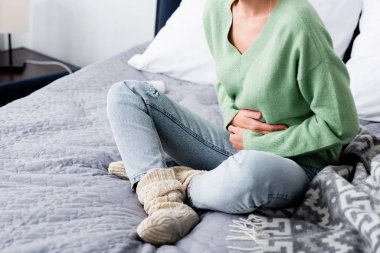 cropped view of woman having abdominal pain and sitting on bed clipart