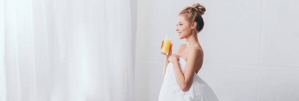 panoramic shot of happy blonde woman with glass of juice in white sheets in bedroom in morning