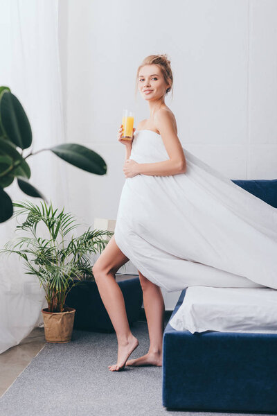 beautiful young woman with glass of juice in white sheets in bedroom in morning