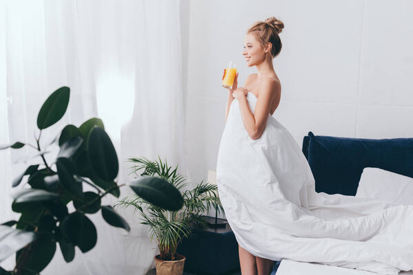 happy girl with glass of juice in white sheets in bedroom in morning