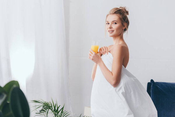 smiling woman with glass of juice in white sheets in bedroom in morning