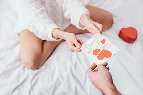 ギフトボックス付きのベッドで男からバレンタインデーのための心を持つグリーティングカードを取って女性のクロップドビュー — ストック写真