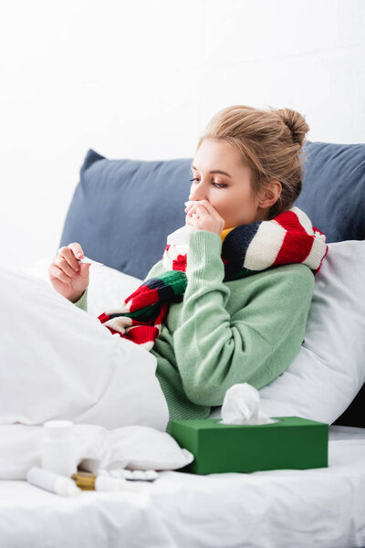 ill woman in scarf having fever and looking at thermometer in bed 