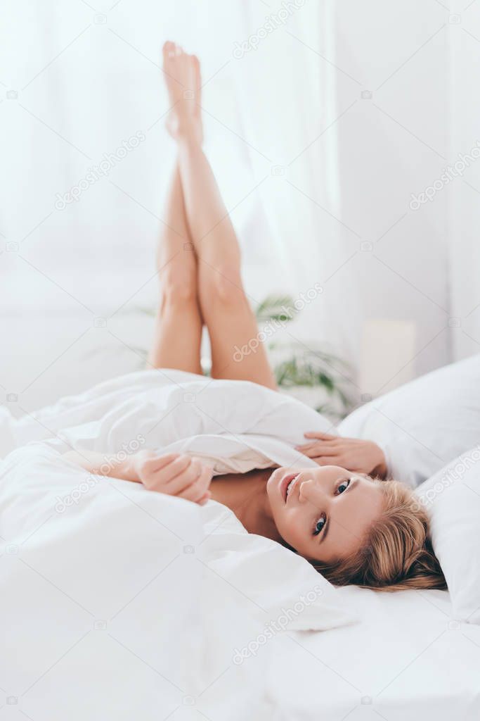 beautiful smiling woman lying with legs up in sheets on bed in morning