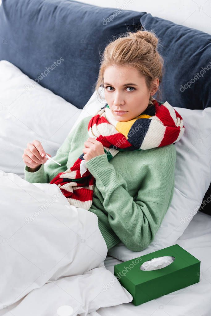 sick woman in scarf having fever and holding thermometer in bed with napkins